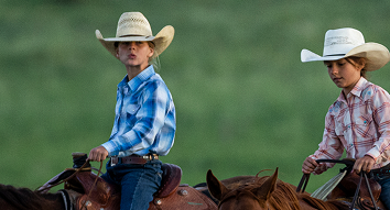Two children wearing cowboy hats and western shirts riding horses outdoors in a grassy field.