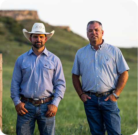 Two men standing outdoors in a grassy field at sunset. The man on the left wears a white cowboy hat, a light blue long-sleeve button-up shirt, and jeans.
