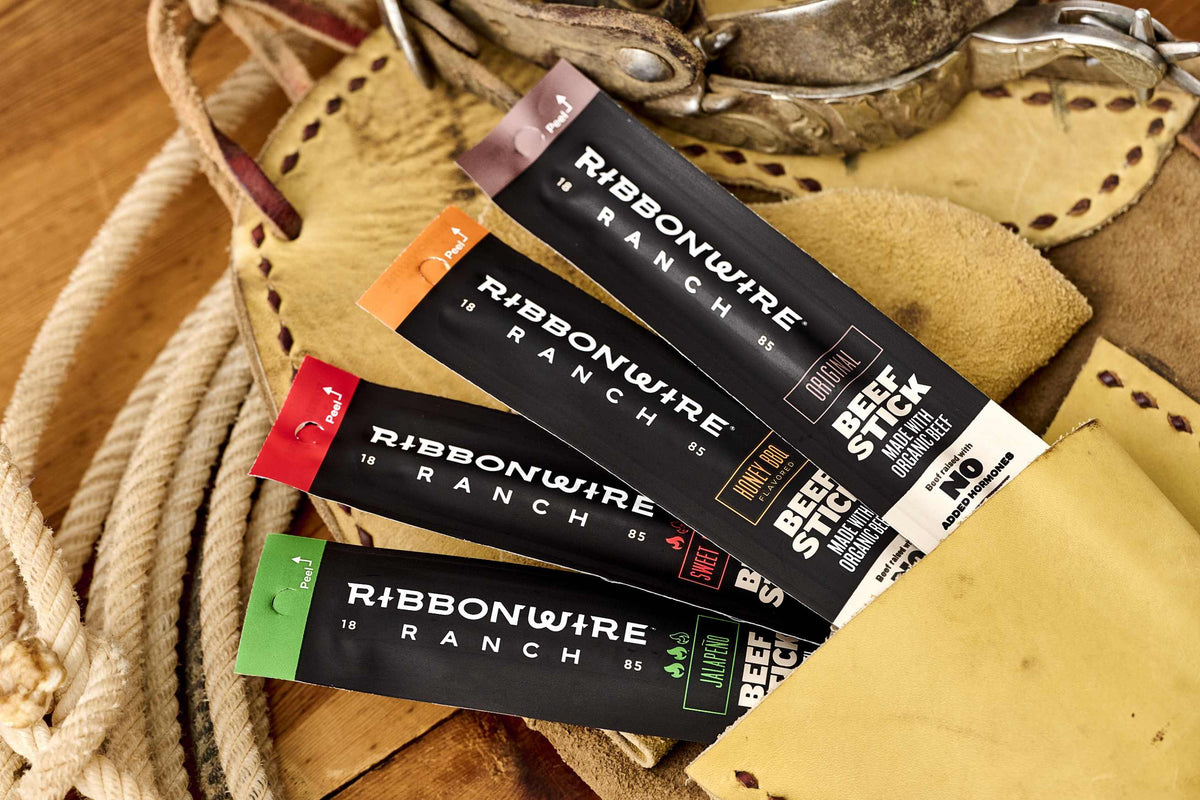 Four packages of Ribbonwire Ranch beef jerky arranged diagonally on a rustic leather and rope surface. The packages are black with different colored tabs (brown, orange, red, green) and white text, with the top package showing 'BBQ BEEF JERKY 12 OZ'.