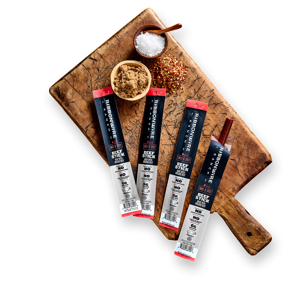 Four packages of Rhinogourmet Beef Stick are arranged on a rustic wooden cutting board with small bowls of salt, spices, and seasoning, photographed from above on a white background.