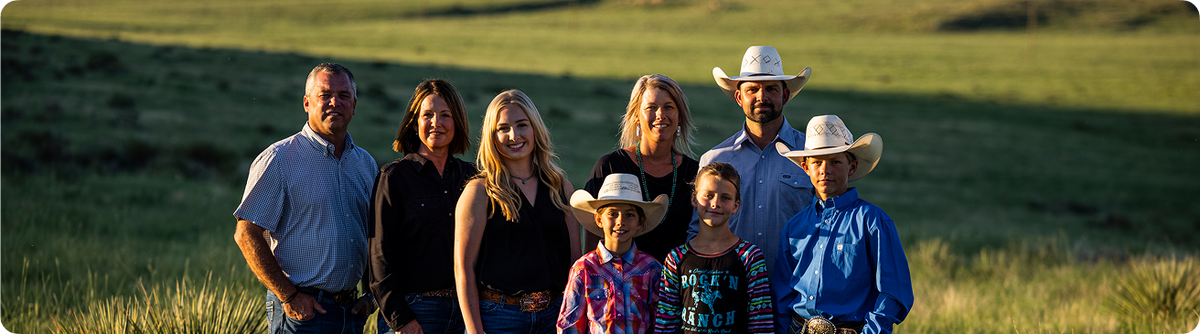 A family of seven posing together outdoors in a grassy field at sunset. The group includes four men and three women, with three wearing cowboy hats.