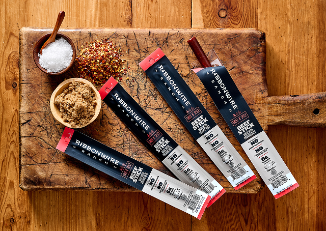 Four packages of Ribbonwire Smokehouse beef sticks arranged on a rustic wooden cutting board with bowls of seasoning and brown sugar on a wooden table.