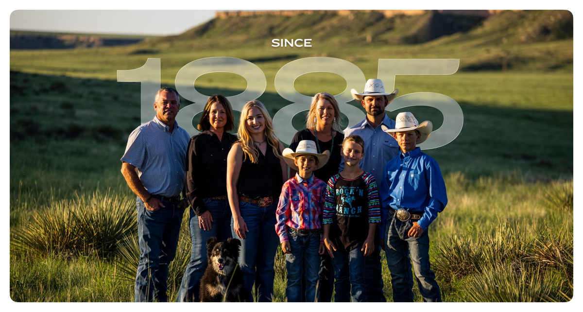 A family portrait of seven people and a dog standing in a grassy outdoor field, with a large semi-transparent 'SINCE 1885' text overlay across the center. The group is dressed in western and casual clothing, with three wearing cowboy hats.
