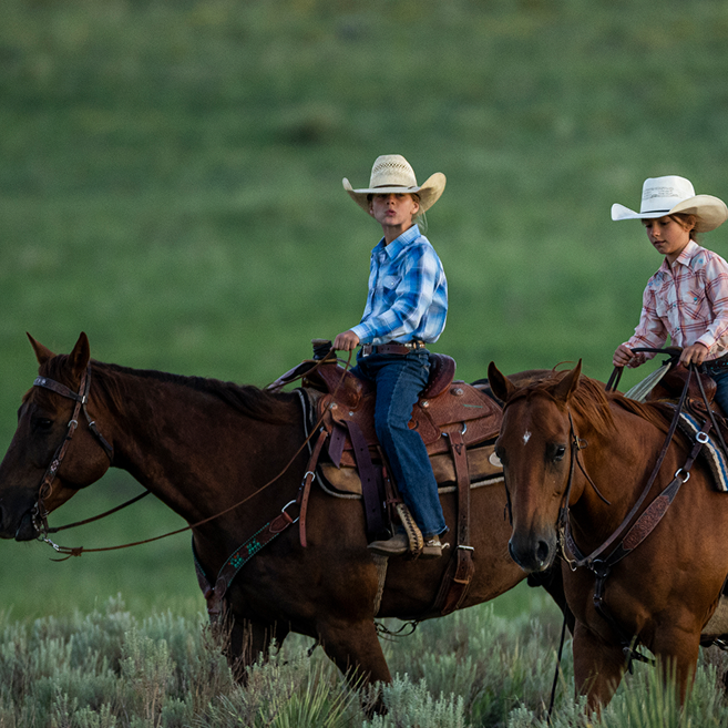 Two young boys wearing cowboy hats and western shirts ride brown horses across a grassy field.