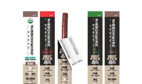 Four Ribbonwire beef sticks packages lined up on a white background, with one stick partially pulled from its wrapper. The packages show different flavor options: Original, Shred & Crisp, Super Sweet, and Zero Sugar.