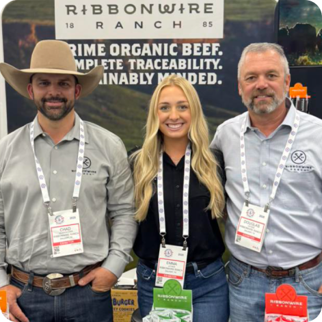 Three people standing together at a trade show booth, wearing branded Ribbons Wire shirts and lanyards, with a large Ribbons Wire Ranch banner behind them.